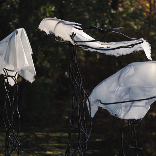 Drei abstrakte Skulpturen, die Quallen ähneln, stehen im Freien. Jede hat einen schwarzen, verdrehten Metallsockel und einen zerfetzten weißen Stoff, der vor einem dunklen, blattreichen Hintergrund drapiert ist.