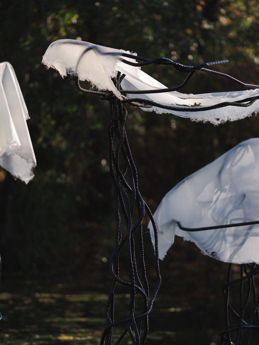 Drei abstrakte Skulpturen, die Quallen ähneln, stehen im Freien. Jede hat einen schwarzen, verdrehten Metallsockel und einen zerfetzten weißen Stoff, der vor einem dunklen, blattreichen Hintergrund drapiert ist.