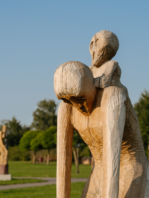 Eine Holzskulptur aus zwei abstrakten menschlichen Figuren, die übereinander sitzen. Die Figuren haben verlängerte Gliedmaßen und vereinfachte Gesichtszüge, die vor einem unscharfen Hintergrund stehen, was dem Bild eine mysteriöse und kontemplative Atmosphäre verleiht.