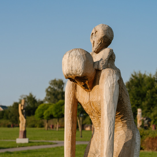 Eine Holzskulptur aus zwei abstrakten menschlichen Figuren, die übereinander sitzen. Die Figuren haben verlängerte Gliedmaßen und vereinfachte Gesichtszüge, die vor einem unscharfen Hintergrund stehen, was dem Bild eine mysteriöse und kontemplative Atmosphäre verleiht.