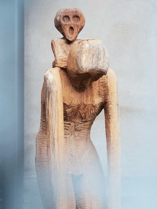 Eine Holzskulptur aus zwei abstrakten menschlichen Figuren, die übereinander sitzen. Die Figuren haben verlängerte Gliedmaßen und vereinfachte Gesichtszüge, die vor einem unscharfen Hintergrund stehen, was dem Bild eine mysteriöse und kontemplative Atmosphäre verleiht.