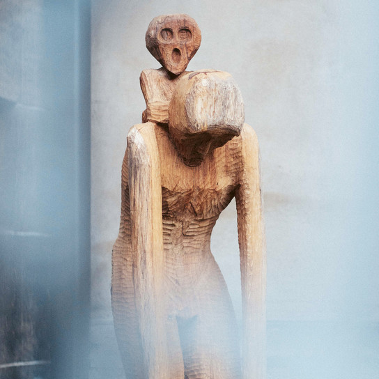 Eine Holzskulptur aus zwei abstrakten menschlichen Figuren, die übereinander sitzen. Die Figuren haben verlängerte Gliedmaßen und vereinfachte Gesichtszüge, die vor einem unscharfen Hintergrund stehen, was dem Bild eine mysteriöse und kontemplative Atmosphäre verleiht.