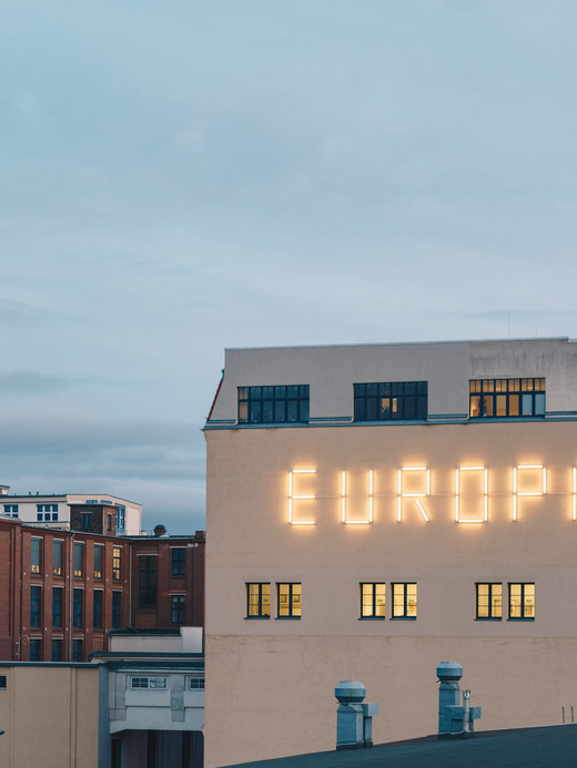 Ein großes Gebäude mit der hellen Aufschrift „EUROPA“ an der Fassade vor einem bewölkten Himmel. Im Hintergrund erhebt sich ein hoher Uhrenturm mit gelben Zifferblättern über die anderen roten Backsteingebäude.