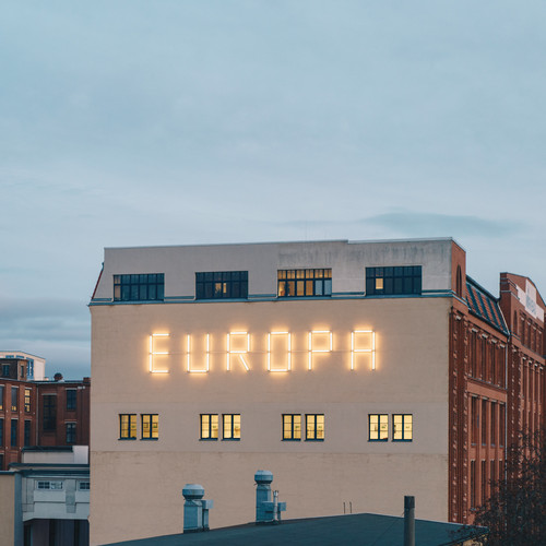 Ein großes Gebäude mit der hellen Aufschrift „EUROPA“ an der Fassade vor einem bewölkten Himmel. Im Hintergrund erhebt sich ein hoher Uhrenturm mit gelben Zifferblättern über die anderen roten Backsteingebäude.