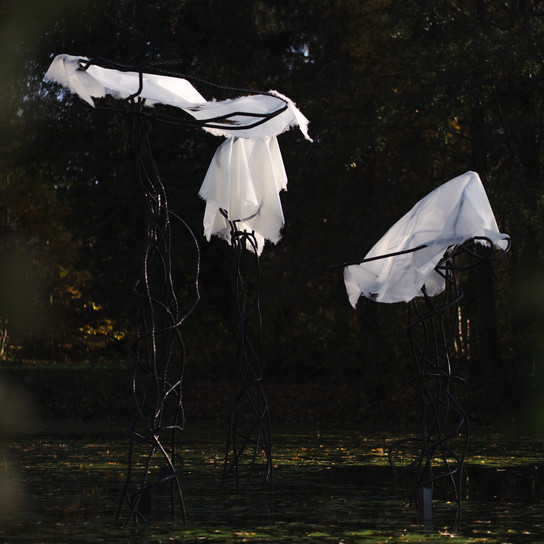 In einem Teich stehen zwei hohe, abstrakte, quallenähnliche Skulpturen aus dunklem, gedrehtem Metall, die mit weißem, lichtdurchlässigem Stoff drapiert sind, umgeben von Bäumen mit Herbstlaub.
