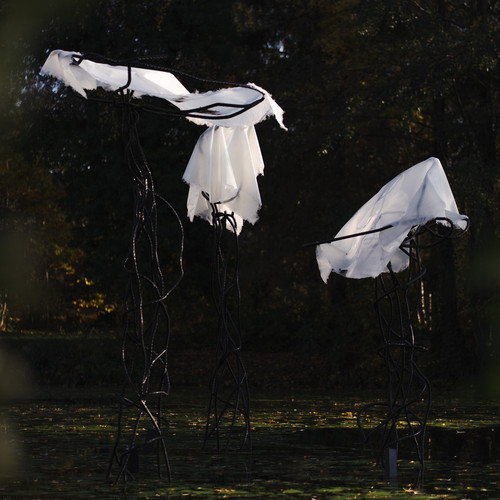 In einem Teich stehen zwei hohe, abstrakte, quallenähnliche Skulpturen aus dunklem, gedrehtem Metall, die mit weißem, lichtdurchlässigem Stoff drapiert sind, umgeben von Bäumen mit Herbstlaub.