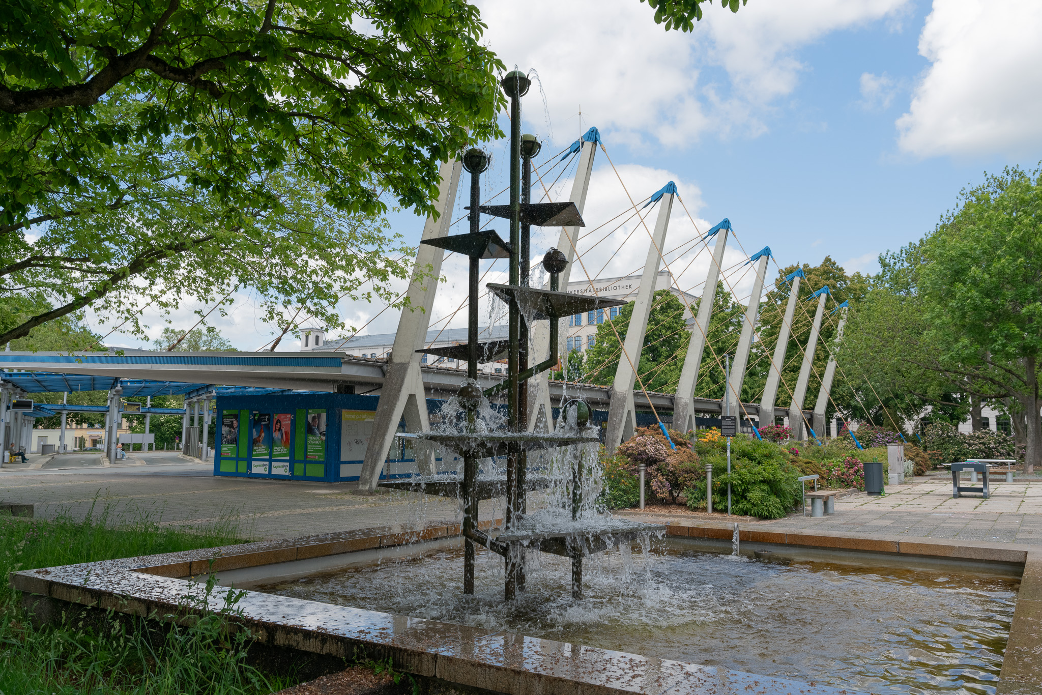 Ein moderner schwarzer Metallbrunnen mit mehreren Stufen steht auf einem Platz und versprüht Wasser in ein flaches Becken. Im Hintergrund befindet sich ein Gebäude mit blauen und weißen Balken, umgeben von Bäumen und Grün.