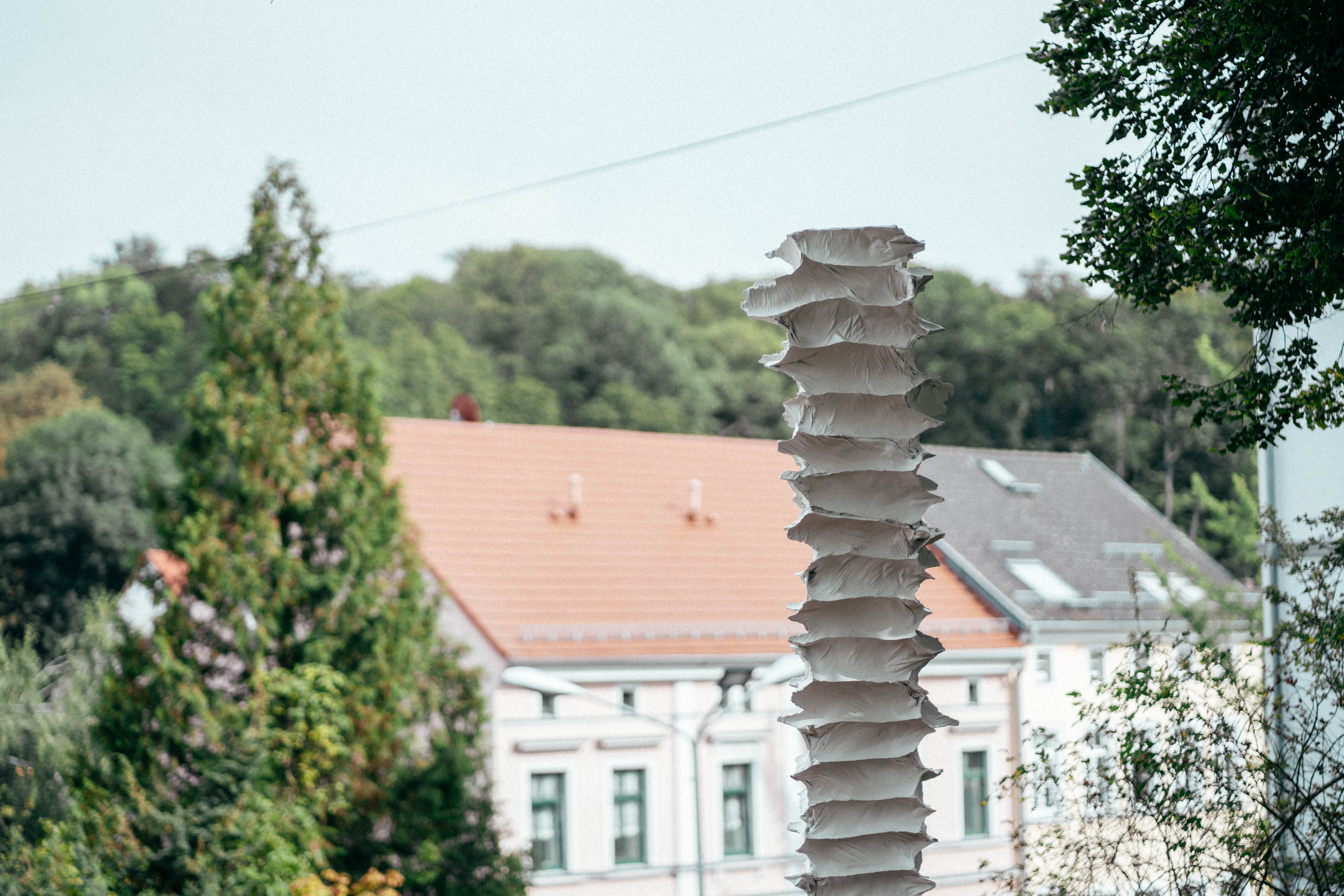 Eine hohe weiße skulpturale Struktur mit gewellten Kanten steht im Freien, mit Bäumen und den Dächern von Wohnhäusern im Hintergrund an einem hellen Tag.