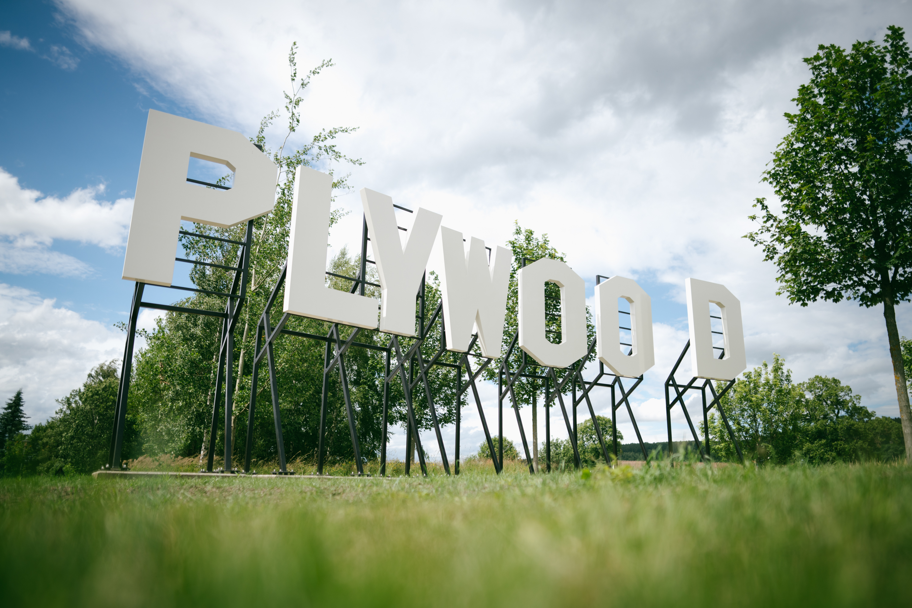 Große weiße Buchstaben mit dem Schriftzug "PLYWOOD" sind auf schwarzen Metallständern im Freien angebracht, umgeben von grünem Gras und Bäumen unter einem bewölkten Himmel.